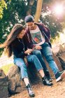 Smiling young couple sitting on rock with smartphone in park — Stock Photo