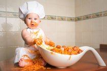 Cheerful little child sitting on tale and having fun while eating pasta from bowl. — Stock Photo