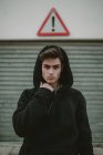 Вдумчивый подросток в черной куртке в капюшоне стоит на улице с восклицательным знаком и смотрит в камеру — стоковое фото