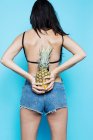 Rear view of woman in bra and denim shorts holding pineapple on blue background — Stock Photo