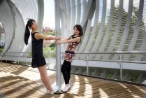 Vue latérale de jolies dames asiatiques souriantes et regardant la caméra tout en se tenant près du poteau métallique sur le pont moderne dans le parc — Photo de stock