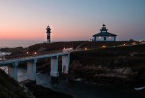 Современный освещенный мост расположен недалеко от маленького островка с маяком и домом в сумерках, Астурия, Испания — стоковое фото