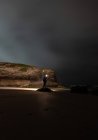 Silhueta de pessoa anônima com lanterna brilhante em pé na costa perto do mar e penhasco na noite nublada, Astúrias, Espanha — Fotografia de Stock