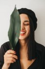 Charmante junge Frau in stylischem Outfit blickt in die Kamera und bedeckt die Brust mit grünem Pflanzenblatt, während sie in der Nähe weißer Wände steht — Stockfoto