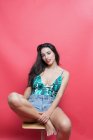 Portrait of young woman in top and denim shorts sitting on chair on pink background — Stock Photo