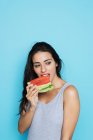 Sinnliche brünette Frau im grauen Body isst frische Wassermelone und schaut vor blauem Hintergrund weg — Stockfoto