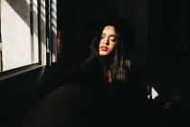 Прекрасна молода жінка тримає очі закритими і підтримує голову, сидячи біля вікна в темній кімнаті і насолоджуючись яскравим сонячним світлом — стокове фото
