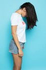 Brunette woman in white shirt and denim shorts with hair covering face standing on blue background — Stock Photo