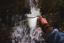 Mano del hombre sosteniendo jarra bajo agua dulce de fuente de agua fría en la naturaleza - foto de stock