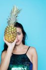 Young woman in glitter top covering eye with pineapple on blue background — Stock Photo