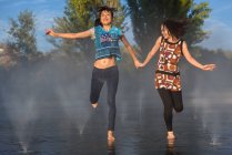 Deux dames asiatiques riant et se tenant la main tout en courant sur l'eau de la fontaine éclaboussante dans le parc — Photo de stock