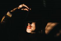 Attraente giovane femmina che copre metà del viso con il cappello mentre in piedi in camera oscura — Foto stock