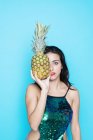 Young woman in glitter top covering eye with pineapple on blue background — Stock Photo