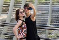 Due affascinanti signore asiatiche sorridenti e in posa per selfie mentre in piedi vicino alla recinzione in metallo nel parco moderno — Foto stock
