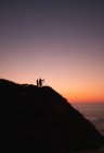 Silhouette of two people with camera on tripod standing on coast near calm sea on background of cloudless sunset sky — Stock Photo