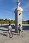 Две молодые азиатки в стильных нарядах улыбаются и катаются на велосипедах возле столба в солнечный день в красивом парке — стоковое фото
