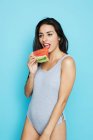 Sensuelle femme brune en body gris mangeant pastèque fraîche et regardant loin sur fond bleu — Photo de stock