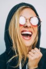 Young blond woman in sunglasses and hoodie laughing on white background — Stock Photo