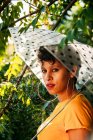 Вид сбоку на привлекательную молодую женщину с зонтиком, смотрящую в камеру, стоя под мокрыми ветвями деревьев в солнечный день в джунглях — стоковое фото