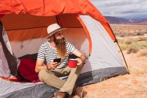 Бородатый парень в повседневной одежде держа кружку горячего напитка и современный смартфон, сидя рядом с палаткой и глядя прочь в красивой природе — стоковое фото