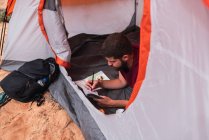 De cima tiro de cara jovem deitado na tenda e desenho em caderno de esboços enquanto acampando no deserto — Fotografia de Stock
