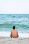 Vue arrière du mâle en short assis sur le rivage sablonneux près de la magnifique mer ondulante à Tylenovo, bulgarie — Photo de stock