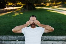 Attractive adult male covering eyes while sitting on bench in park on sunny day — Stock Photo