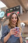 From below shot of attractive young female smiling and browsing modern smartphone while standing on blurred background of railway station — Stock Photo