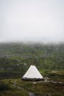 Белая палатка на зеленой земле у подножия горы, покрытая густым белым туманом — стоковое фото