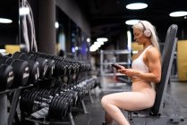 Спортсменка, сидящая в спортзале с мобильным телефоном и наушниками — стоковое фото