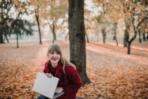 Young lady in red coat using device and sitting on seat in autumn forest — Stock Photo