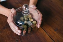 From above shot of crop hands covering stacks of coins with glass lid on wooden table — Stock Photo