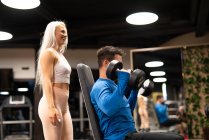 Mujer ayudando al hombre a hacer ejercicio con pesas en el gimnasio - foto de stock