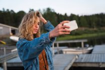 Junge attraktive Frau mit blonden Haaren in Jeansjacke steht am Ufer des Flusses und macht Selfie mit Handy — Stockfoto