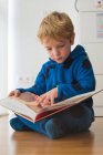Concentré blonde garçon lecture livre sur plancher en bois — Photo de stock