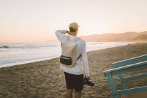 На задньому плані молодий хлопець з професійною фотокамерою йде по піщаному пляжу під час заходу сонця в Санта-Моніці, каліфорнія. — стокове фото