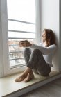 De cima tiro de fêmea adulta atraente em suéter de malha segurando caneca de bebida quente e olhando para a rua enquanto sentado na soleira da janela no quarto aconchegante — Fotografia de Stock