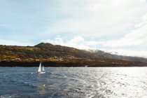 Вид одиночного корабля с белыми салями, плывущими в синей морской воде на фоне зеленых гор при солнечном свете — стоковое фото