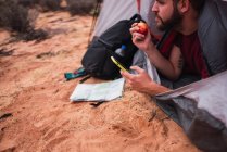 Урожай бородатий чоловік їсть свіже яблуко і переглядає сучасний смартфон, лежачи в наметі під час кемпінгу в пустелі — стокове фото