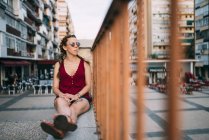 Thoughtful red-haired girl with braids and sunglasses sitting near railing in city — Stock Photo