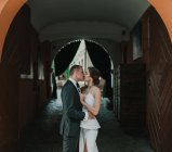 Vue latérale de la jeune mariée et le balai embrassant et s'embrassant tout en se tenant dans l'arche de vieux bâtiment sur la rue de la ville — Photo de stock