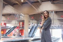 Вид збоку позитивної леді в елегантному вбранні посміхається і дивиться на камеру, стоячи на сучасному залізничному вокзалі — стокове фото