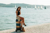 Trendy woman in high heels sitting on grungy stub on paved seafront with sailing boats on background — Stock Photo