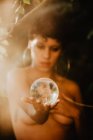 Jovem morena topless cobrindo peito e segurando bola transparente de vidro em madeiras verdes — Fotografia de Stock