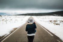 Vue arrière de la jeune personne en tenue élégante marchant au milieu de la route asphaltée par une journée d'hiver nuageuse dans une belle campagne — Photo de stock