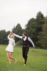 Trendy adult groom and bride in elegant suits holding hands and jumping with excitement on green meadow — Stock Photo