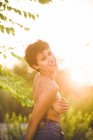 Confident provocative woman in denim jeans standing topless covering breast and looking at camera in nature — Stock Photo