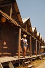 Unrecognizable fit tanned female with brown hair and straw hat wearing jean shorts and white top standing and leaning on wooden building at beach — Stock Photo