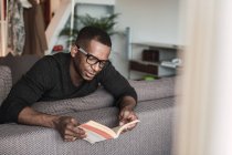 Fokussierter afrikanisch-amerikanischer Mann mit Brille liest Buch, während er zu Hause auf dem Sofa sitzt — Stockfoto