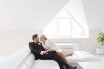 Vue des jeunes mariés modernes élégants dans l'intérieur blanc simple embrassant sur le canapé dans la lumière du jour douce — Photo de stock